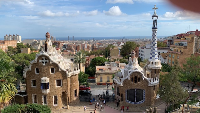 Two characteristic stone buildings with white decorative tops in a park, with a city in the distance under a blue sky with clouds.