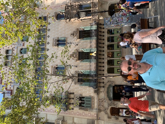 A historical building with ornate balconies and colourful tiles is partly hidden by leafy trees, while visitors gather on the square in front of the building.