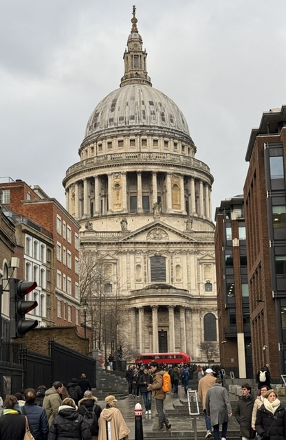 The dome of St Paul's Cathedral at the end of a street, with a red double-decker bus and people on the steps under a grey sky.