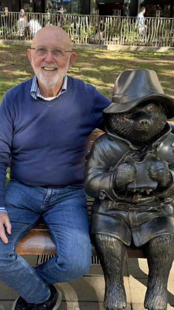 A man on a bench beside the bronze statue of a small bear in a hat and coat, in the sunshine.