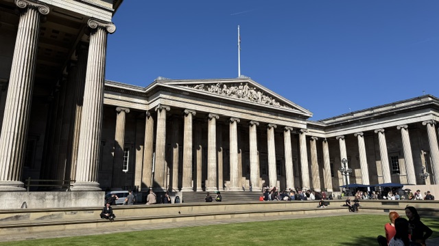 The colonnade of the British Museum with its triangular pediment, people sitting on the steps under a clear blue sky.