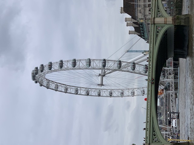 A large observation wheel across the Thames, with the green railing of a bridge in the foreground under cloud.