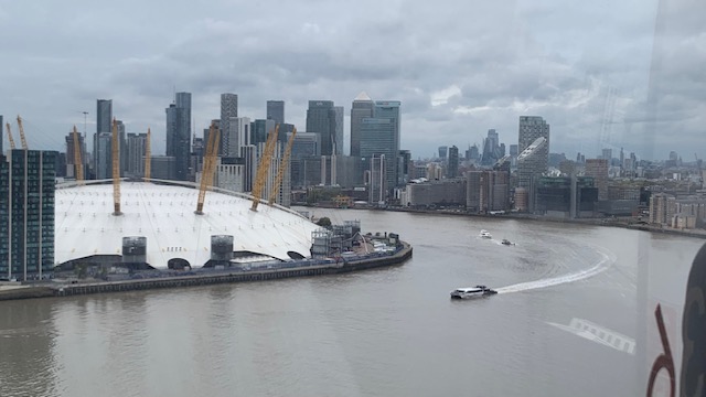 A white dome with yellow masts at the water's edge, with tall office towers behind it and boats on the river.