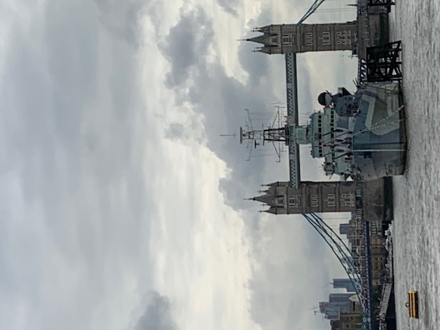 A grey warship moored in the river with Tower Bridge behind it, under a heavy overcast sky.