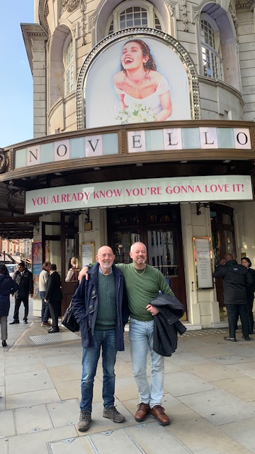 Two men outside the entrance of a theatre, under the illuminated sign above the doors.