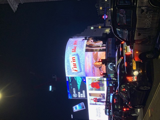 Lit advertising screens above a junction at night, with taxis and cars waiting in front of them.