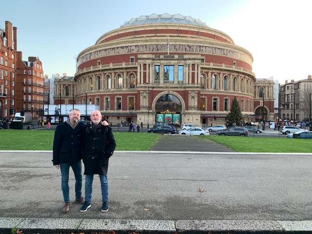 Two men in the street in front of a round brick concert hall with its wide frieze, in the early evening.