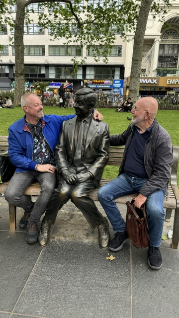 Two men on a bench on either side of a bronze statue of a seated man, in a small park.