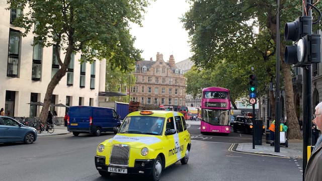 A yellow taxi and a pink double-decker bus at a junction, with trees and a brick facade behind.