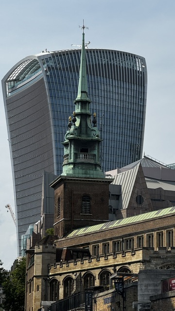 A green church tower with a spire, with the curved glass facade of a modern office tower behind it.