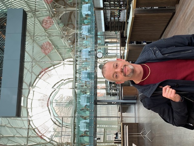A man in a foyer under a high glass roof with iron arches, tables on the floor above behind him.