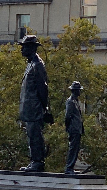 Two bronze statues of men in hats and suits, high on a plinth in front of a tree.
