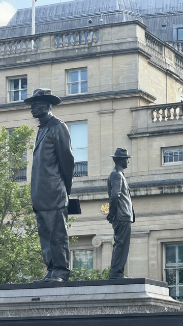 The same two bronze men in hats, now with the pale facade of a large building behind them.