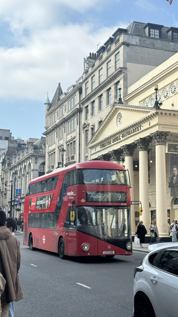 A red double-decker bus in front of a columned theatre, in a busy shopping street.