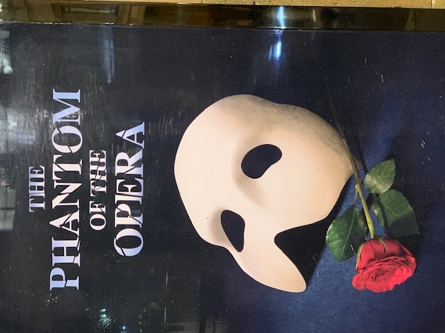 The poster for The Phantom of the Opera with its white mask and a red rose, behind glass.
