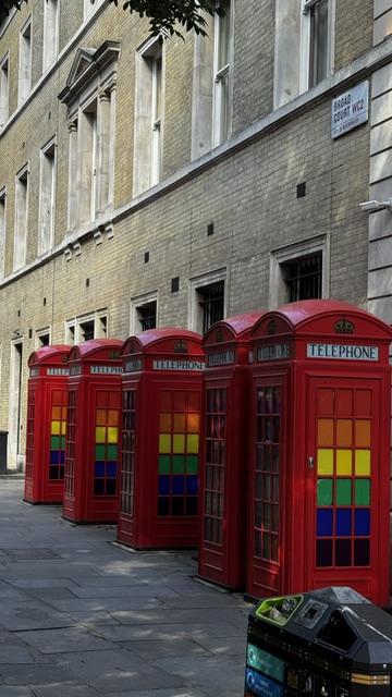 Five red telephone boxes in a row with panes in rainbow colours, along a pale stone facade.