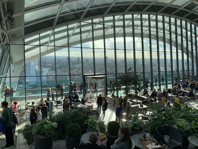 A large glass conservatory high in a building, with plants, seating and a view over the city.