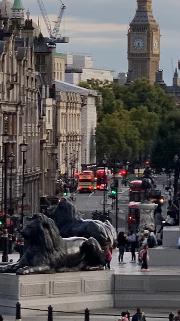 A bronze lion in the foreground, with a street full of red buses and a clock tower beyond.