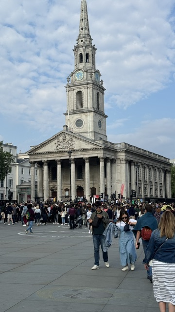 A white church with columns and a tall spire at the edge of a square, with many people in front of it.