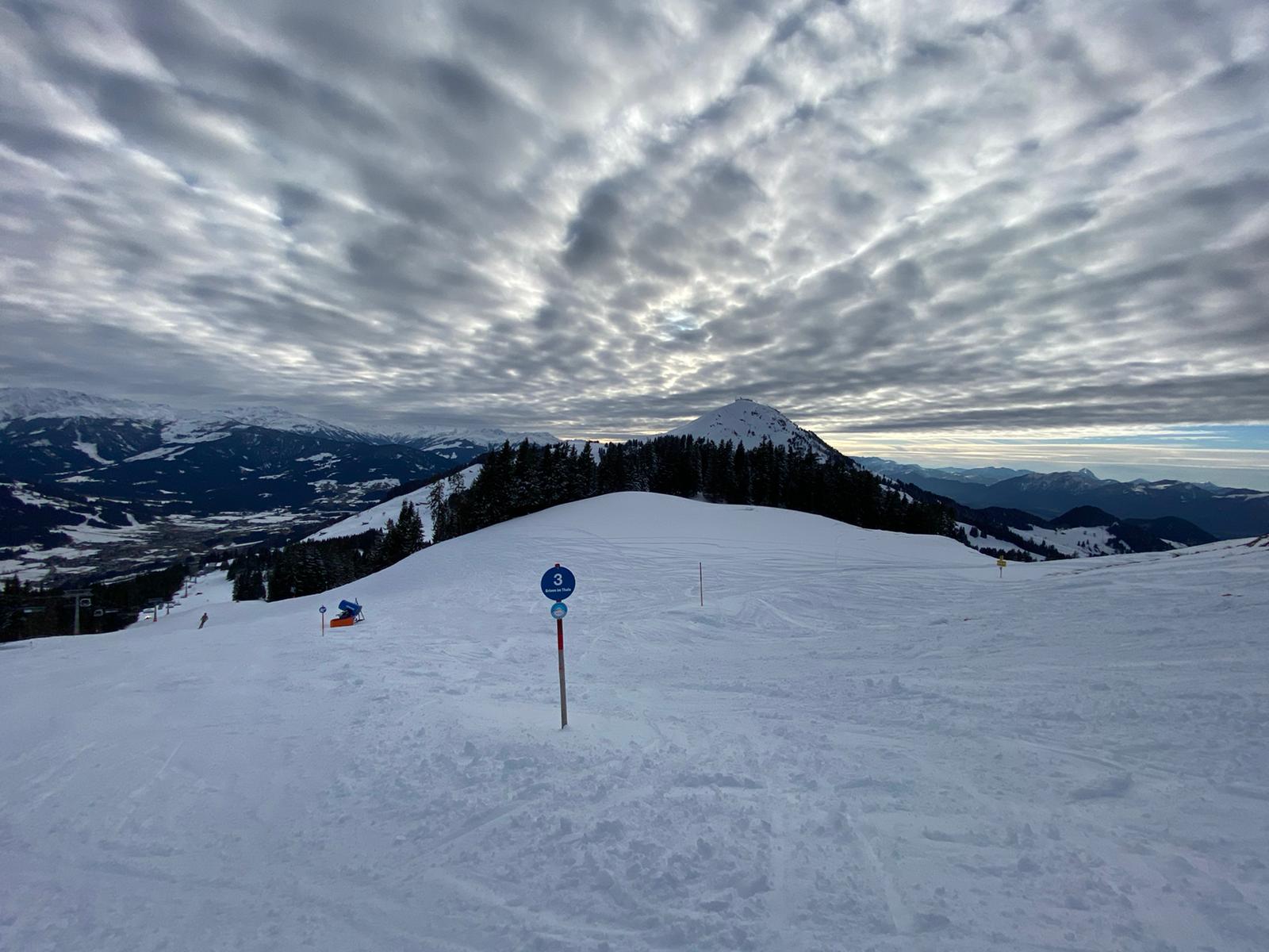 A wide field of snow on a mountain plateau, with peaks in the distance and a cloudy sky above.