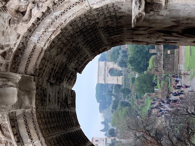Through the curved span of the Arch of Titus, a view of the Forum Romanum, with the Arch of Septimius Severus beyond it among the ruins and the pines.
