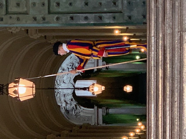 A Swiss Guard in a colourful uniform keeps watch at a lit hall with vaulted arches.