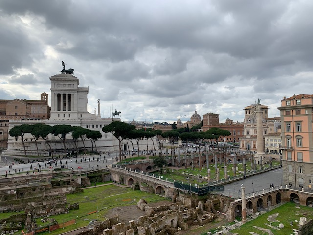 View over the ruins of the Forum of Trajan, with the white Monumento a Vittorio Emanuele II in the background under a heavily clouded sky.