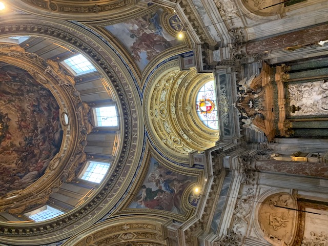 A richly decorated dome with frescoes and gold-coloured ornaments, with below it a stained-glass window above the altar of a church.