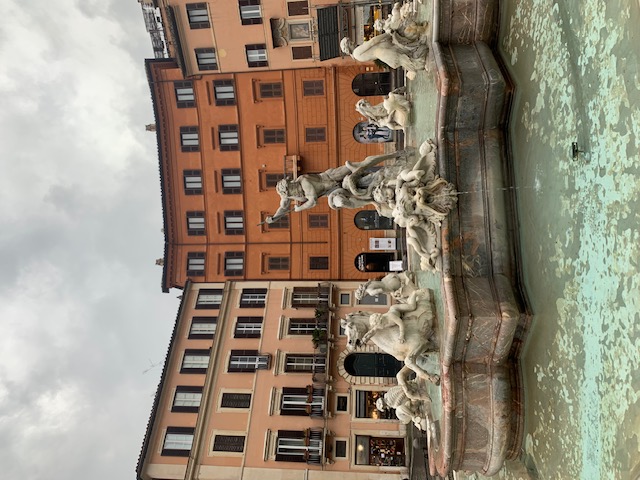 The Fontana del Nettuno on Piazza Navona, with its statues of seahorses and tritons, against the coloured facades under a cloudy sky.