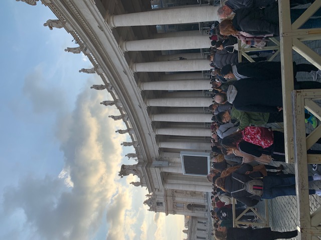 Rows of people wait at the colonnade of St Peter's Square under a cloudy sky with some evening light.