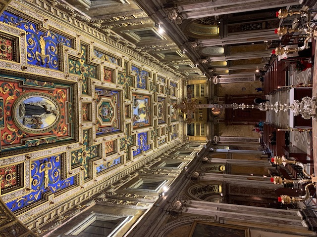 A richly gilded, colorful coffered ceiling towers above the nave of this church, with marble columns and pews that face the altar.