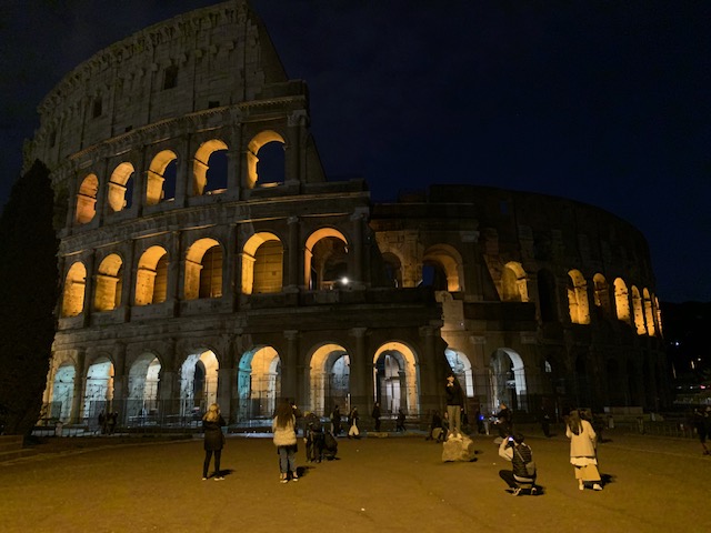 The Colosseum bathes in warm light against the dark evening sky, while visitors walk around in front of it and pose.