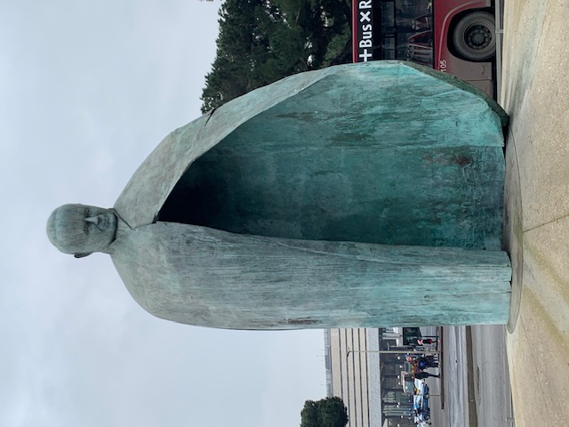 A green weathered bronze statue with a hollow, cloak-shaped form dominates the square, with a bus stop and an overcast sky in the background.