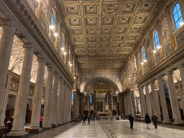 The gilded coffered ceiling and rows of marble columns of the Santa Maria Maggiore lead the eye towards the illuminated altar in the apse.