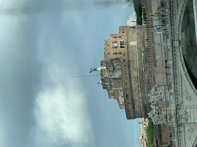 Castel Sant'Angelo rises up behind the decorated bridge arch, under a cloudy sky.