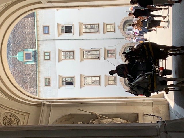 A two-horse Fiaker carriage passing through a tall archway of the Hofburg into a sunlit courtyard.