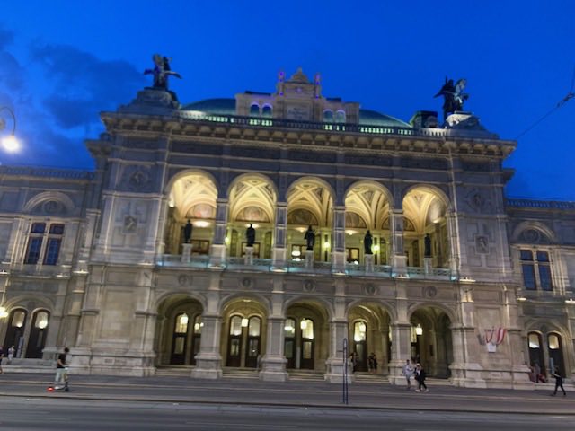 The Vienna State Opera lit warm against a deep blue evening sky, its arcaded loggia glowing.