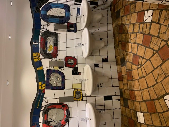 A public washroom tiled in Hundertwasser's style: irregular white tiles, coloured mosaic mirrors, an uneven brick floor.